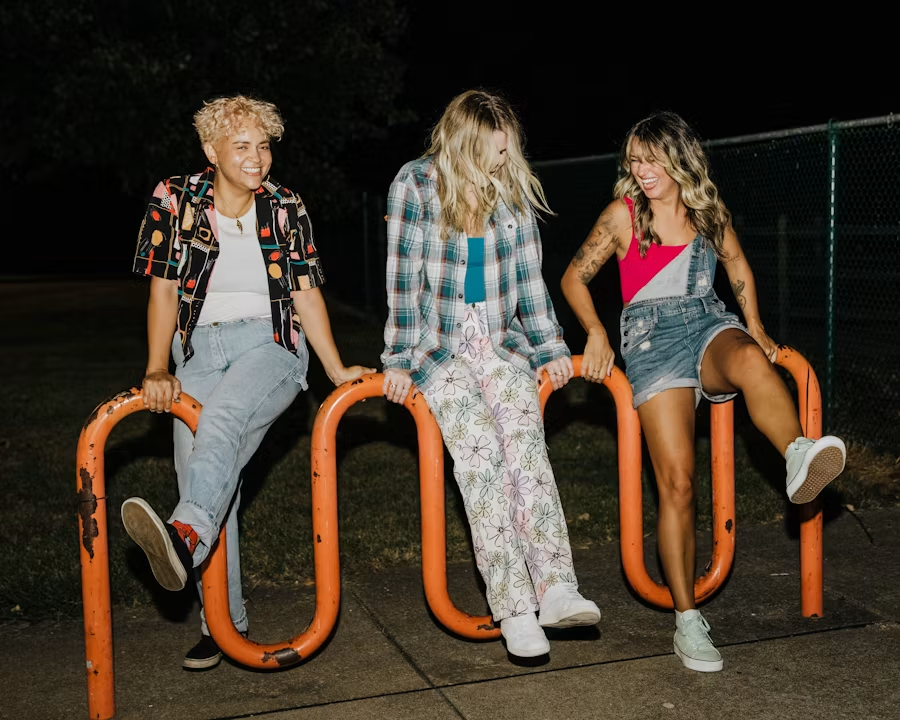 Three friends laughing and posing on an orange looping bike rack at night.