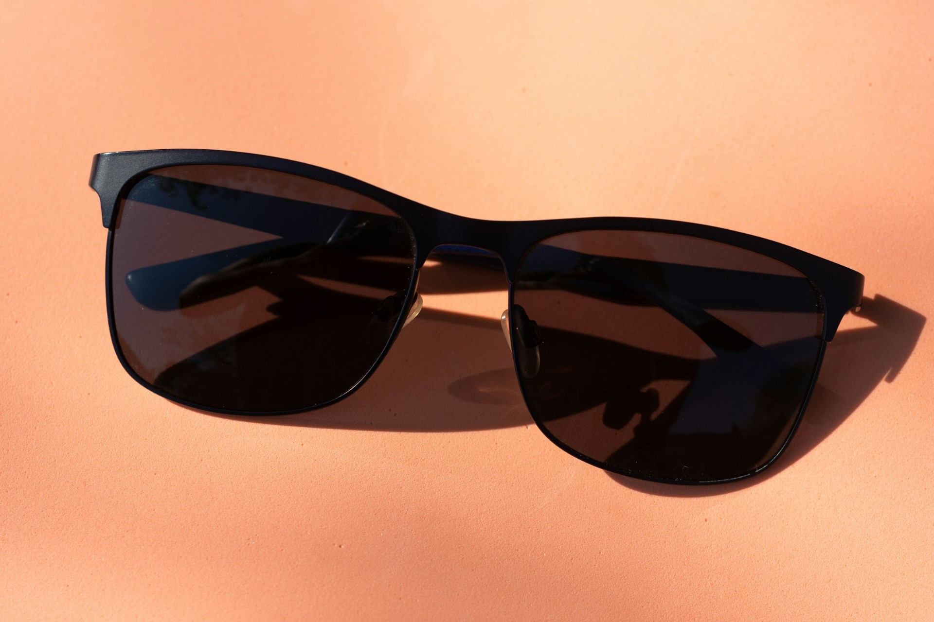 A pair of dark sunglasses resting on a warm peach surface, catching the sunlight.