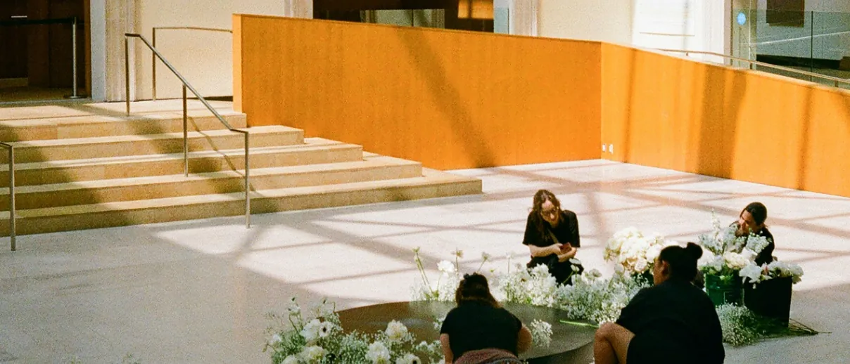 People arranging white flowers around a low circular plinth in a sunlit museum atrium with tall arched windows.